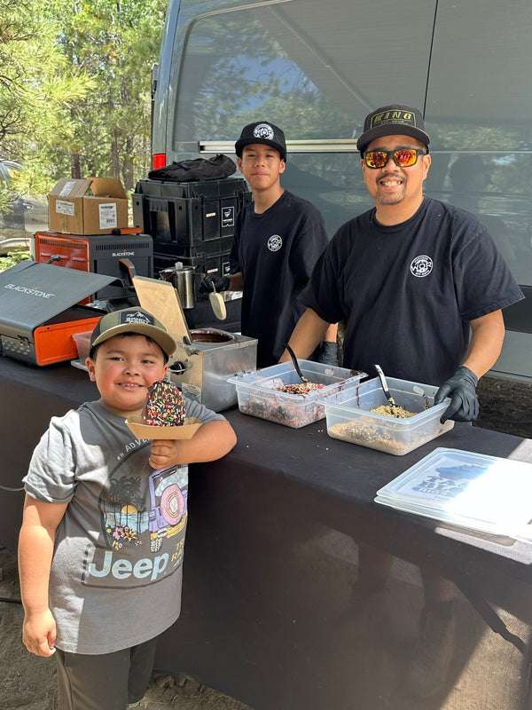 A smiling child holding a chocolate-covered ice cream cone with sprinkles while two volunteers prepare toppings at a treat station during the Big Bear Experience 2026 overlanding event.
