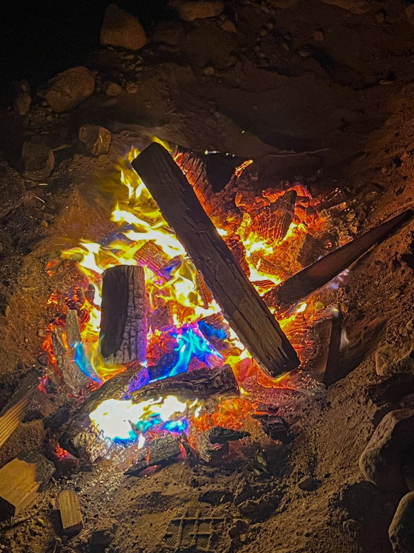 Close-up photo of a glowing campfire during the WOR DCP Giveback event, with vivid blue, green, and yellow flames burning around charred logs at night in the desert.