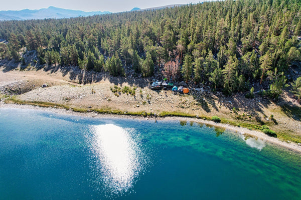 Aerial view of rooftop tents and vehicles camped along a pristine alpine lake surrounded by dense pine forest during the Coyote Flats overland trip, September 4–7, 2026.