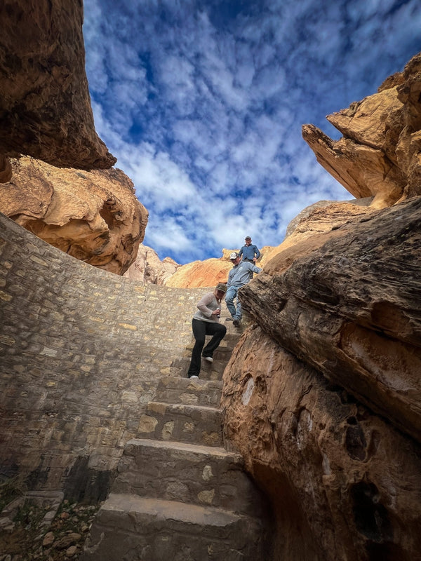 Group of people hiking down carved stone steps inside a narrow slot canyon near the Double Negative land art site during a Weekend Offroad Nevada overlanding adventure, surrounded by rugged red rock walls and dramatic cloud-filled sky.