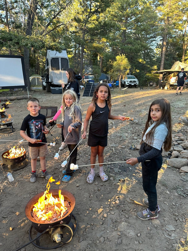 Four children roasting marshmallows over portable fire pits at the Big Bear Experience 2026, surrounded by overlanding campsites, tents, and vehicles in a forest setting.