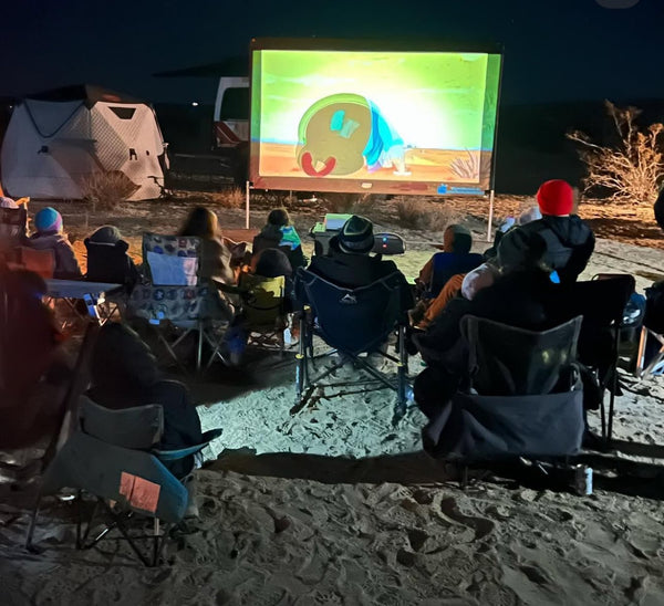 A group of adults and children sit in camping chairs on desert sand watching a movie projected on a large outdoor screen during the WOR DCP Giveback event at night.