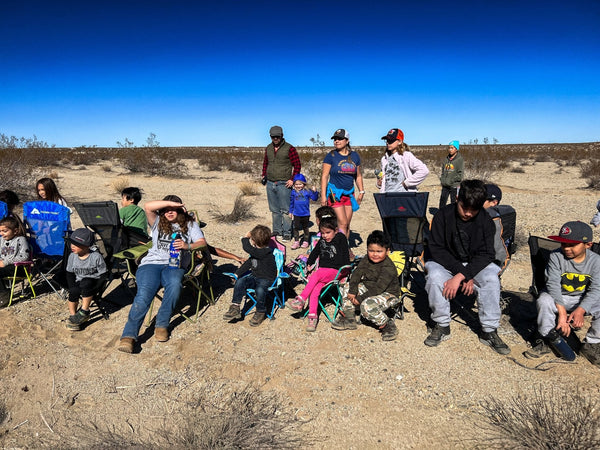 A group of kids sitting in camping chairs in the desert during the WOR DCP Giveback event in January 2026. Bright blue sky, off-road families gathered together enjoying outdoor time.