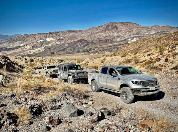 Three off-road vehicles driving in a dusty convoy through rocky desert terrain with mountains in the background during the Weekend Offroad Death Valley trip.