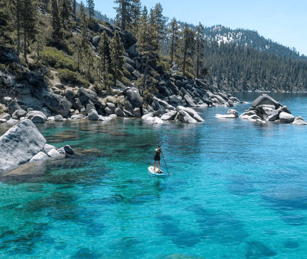 Person paddleboarding on crystal clear blue water surrounded by boulders and pine trees at Lake Tahoe during a Weekend Offroad trip.