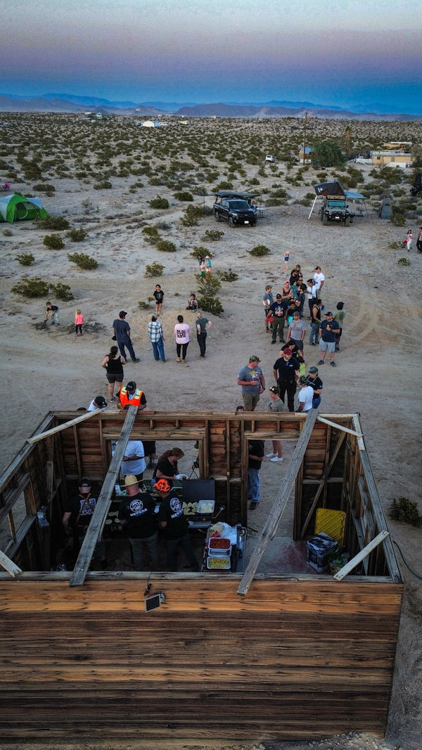 Aerial view of families lined up for dinner at an old wooden cabin structure during the WOR DCP Giveback event in January 2026, surrounded by desert landscape, tents, and off-road vehicles at sunset.