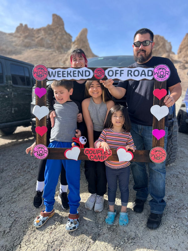 A smiling family poses with a decorated Weekend Off Road couples camp photo frame during the Valentines Experience event, with rugged desert rock formations in the background.