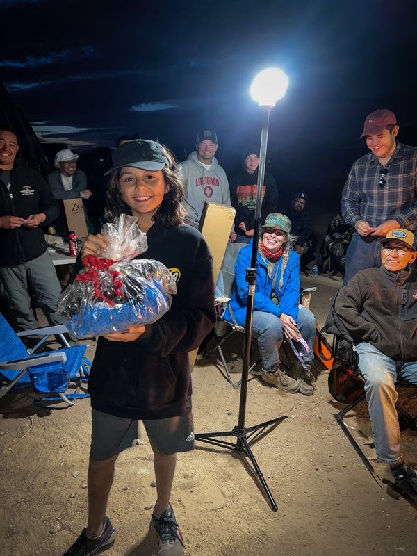 A smiling child holds a wrapped raffle prize during the WOR DCP Giveback event, surrounded by happy attendees sitting in camp chairs under a bright light in the desert at night.