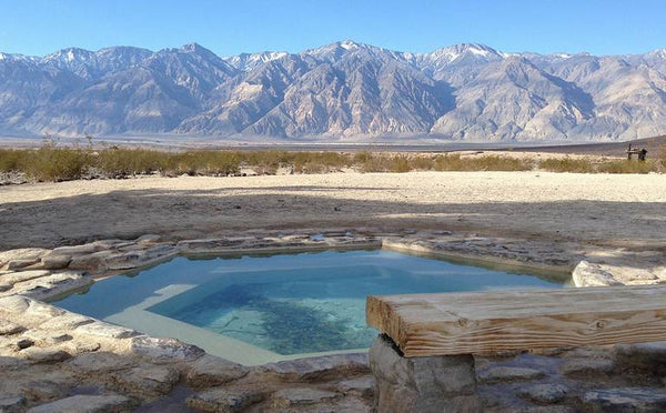 A natural stone hot spring pool in the desert with a wooden sitting ledge, set against a vast backdrop of rugged snow-dusted mountains under a clear blue sky.