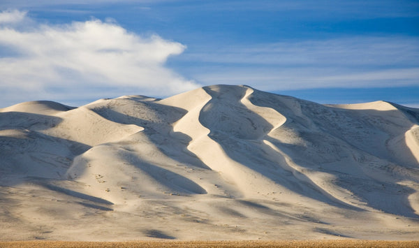 Large rolling sand dunes in bright daylight with soft shadows and a blue sky above, resembling the desert landscape of Death Valley.