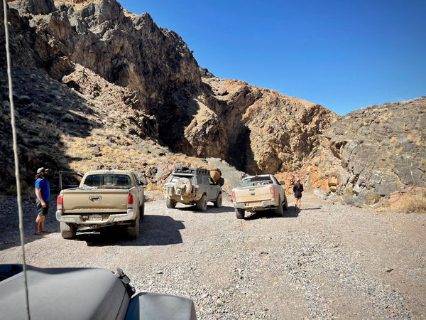 Group of dusty off-road trucks parked along a rocky canyon trail in Death Valley, with people standing near the vehicles under a bright blue sky.