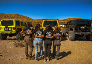 Group of women wearing Weekend Offroad shirts with their arms around each other, standing in front of off-road Jeeps in the desert.